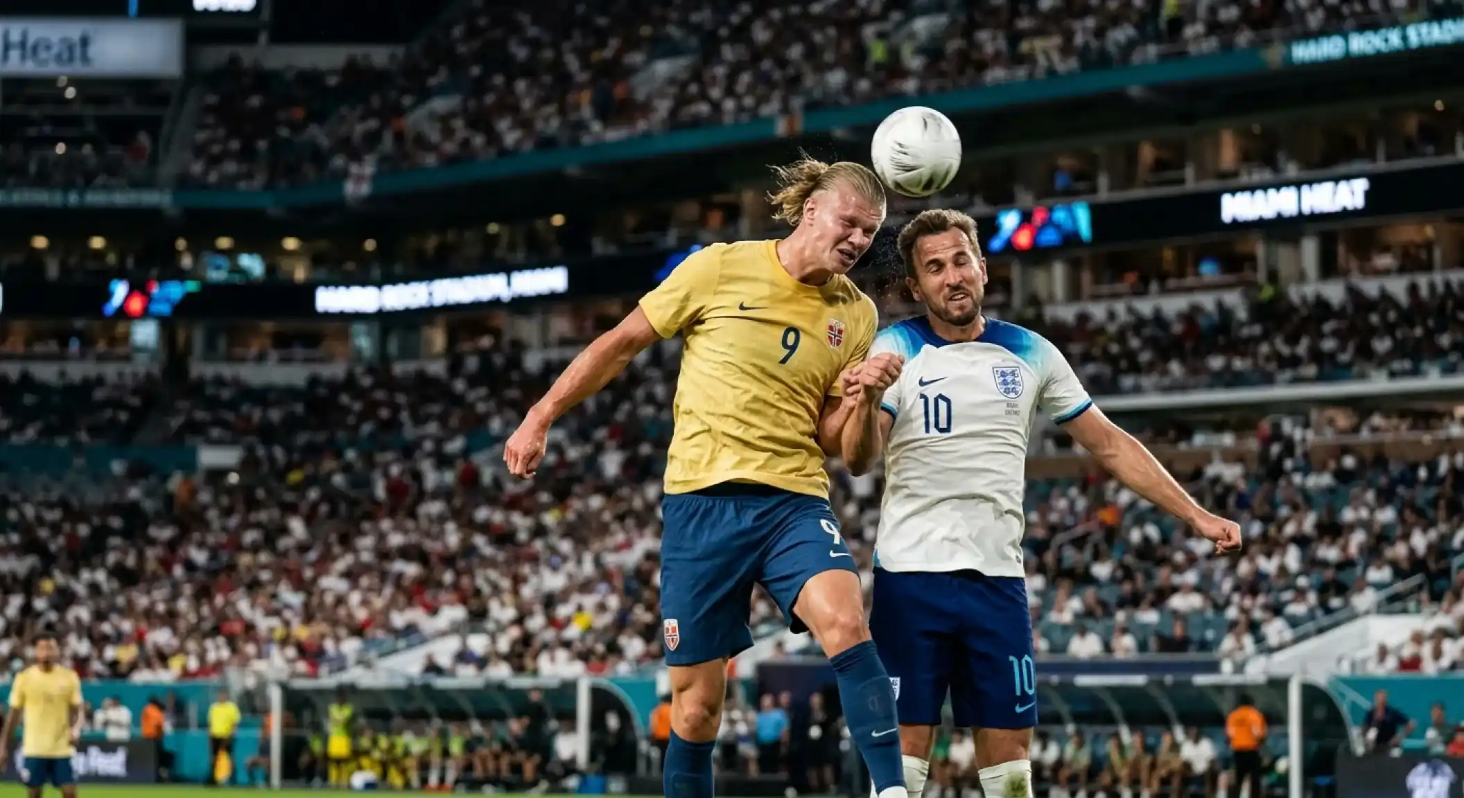 Angleški in norveški nogometaš v borbi za žogo v dežju pod reflektorji Hard Rock Stadium, na ozadju mavrične tribune